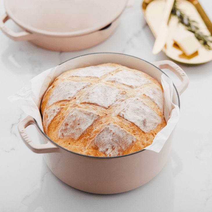 Freshly baked bread in a pink Dutch oven on a marble countertop, with butter and herbs on a tray in the background.