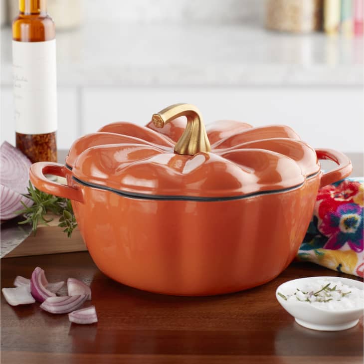 Orange pumpkin-shaped casserole dish with a gold stem handle on a wooden countertop, surrounded by sliced onions and herbs.
