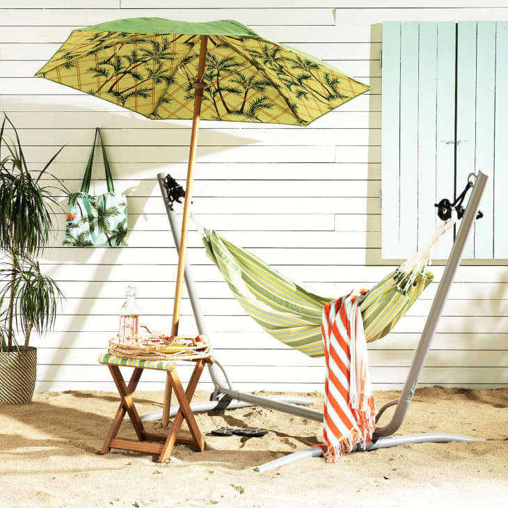 Hammock with green and white stripes under a palm-patterned umbrella, next to a wooden table with drinks on sandy ground.