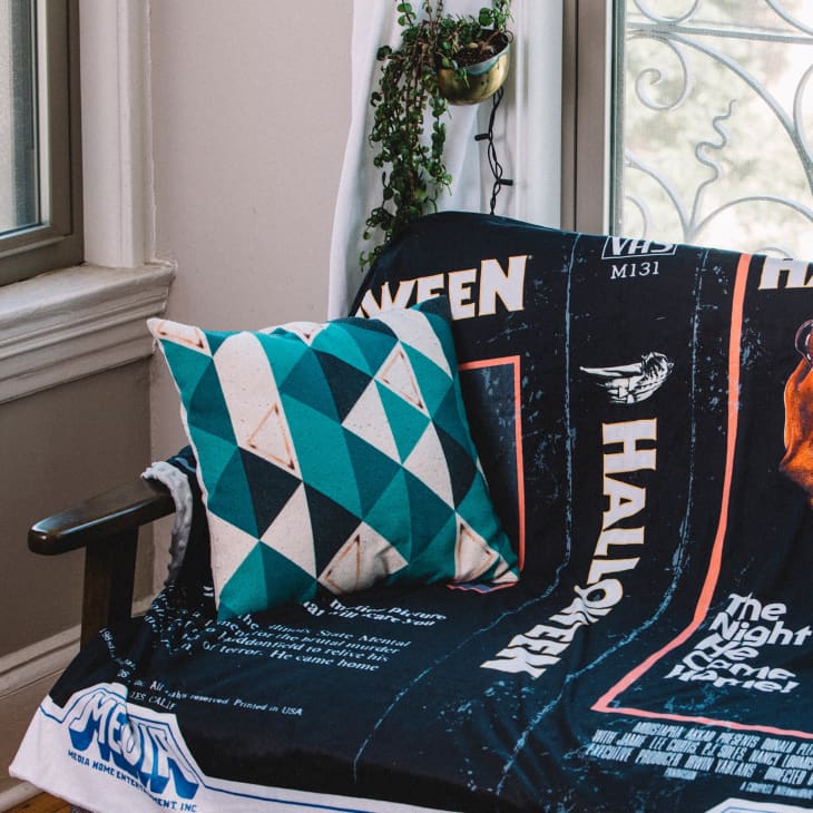 Teal geometric pillow on a chair with a Halloween-themed blanket, near a window with a hanging plant.