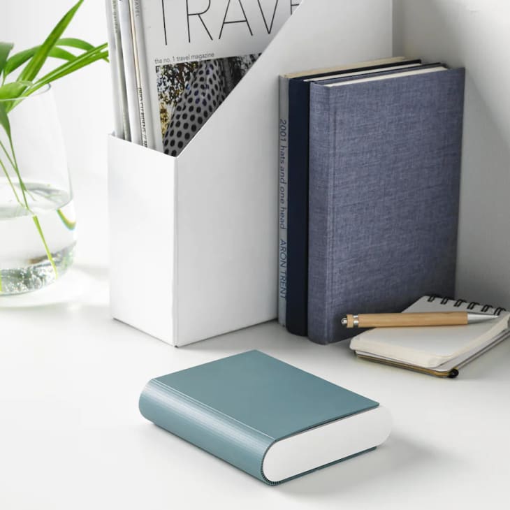 Books and magazines in a white holder, with a blue book, notepad, and pen on a desk.