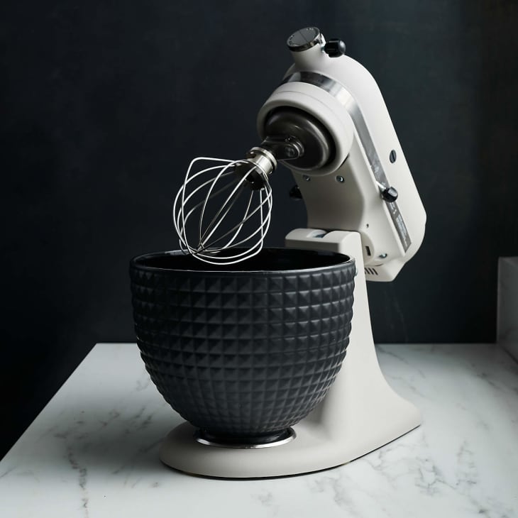 Stand mixer with a wire whisk attachment and textured black mixing bowl on a marble countertop.