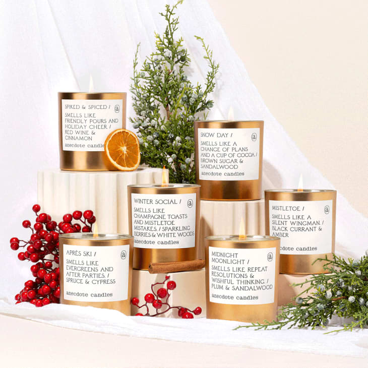 Holiday-themed candles with labels, surrounded by red berries, greenery, and a dried orange slice.