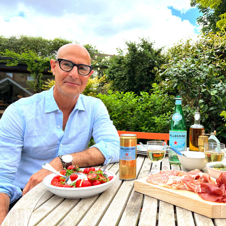 Man in blue shirt sitting at outdoor table with salad, charcuterie, and drinks, surrounded by greenery.