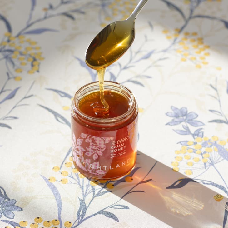 Jar of Brightland Kauai Honey with a spoon drizzling honey, on a floral-patterned surface.