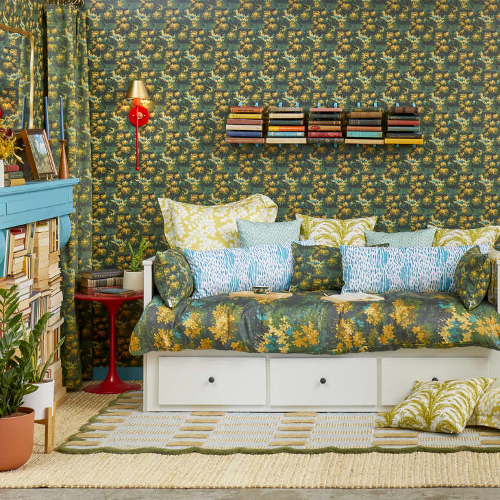 Head on view of a white day bed, in a room with green and yellow floral wallpaper.  The bed is made with a large assortment of pillows with different prints.
