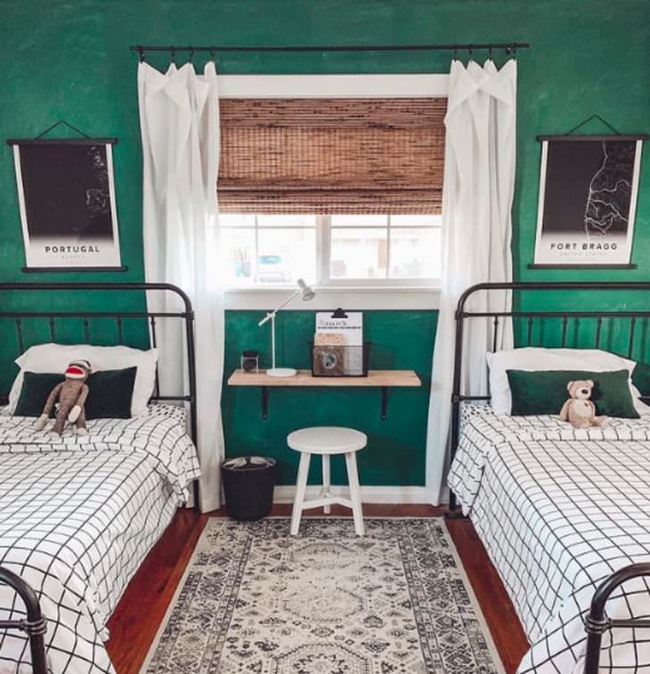 Two twin beds with checkered bedding, green walls, and wall art. A small shelf with books and a lamp is between the beds.