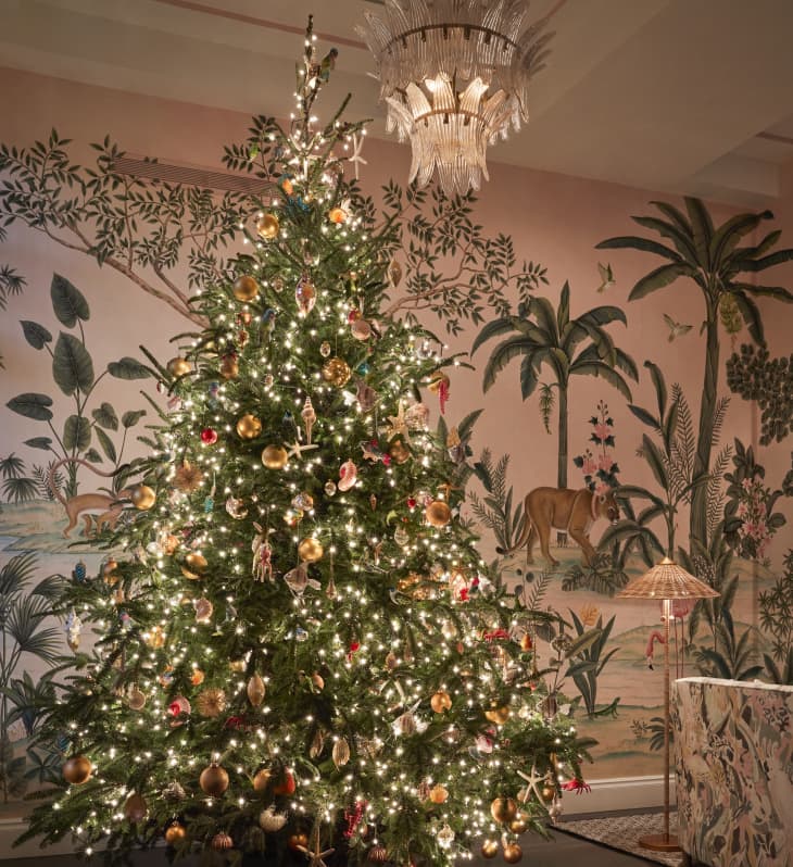 Christmas tree with gold and red ornaments, lit with white lights, in a room with tropical-themed wallpaper and chandelier.