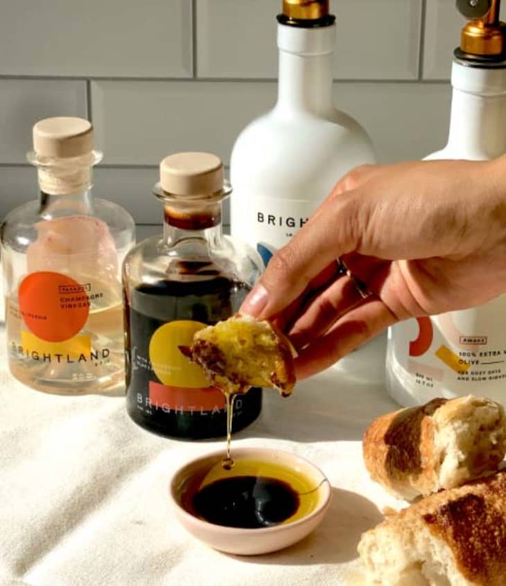 Hand dipping bread in olive oil and vinegar with Brightland bottles in the background.