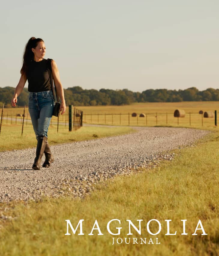 Woman walking on a gravel path through a field, wearing jeans and boots, with hay bales in the background.