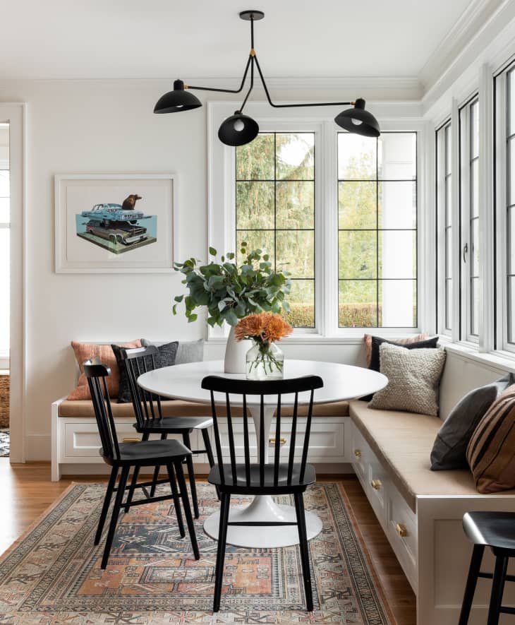 Dining nook with round white table, black chairs, built-in bench, and large windows. Vase with flowers on table.