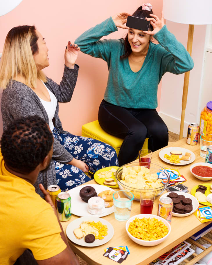 Three friends playing a game with a phone, surrounded by snacks and drinks on a table.