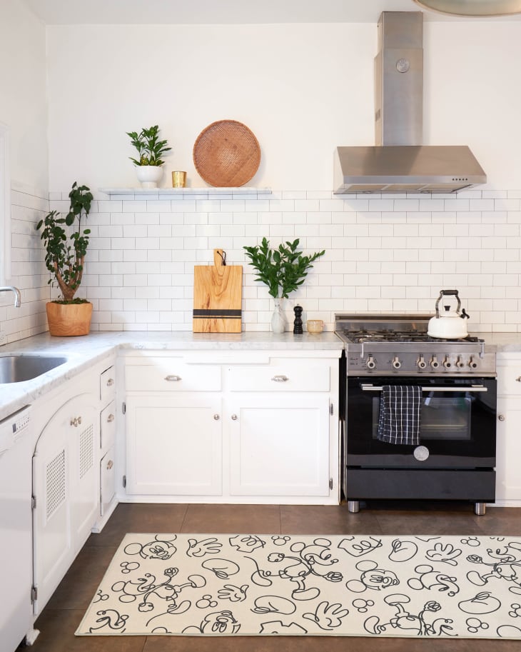 Modern kitchen with white cabinets, subway tiles, black stove, plants, and a Mickey Mouse patterned rug.