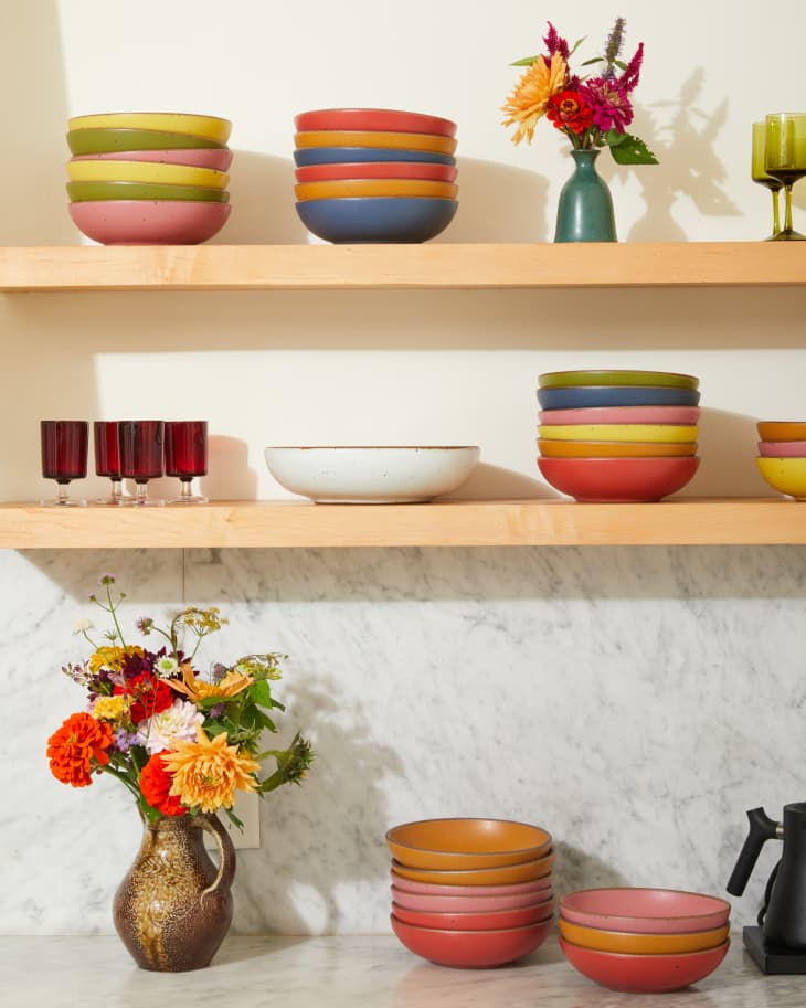 Colorful ceramic bowls stacked on wooden shelves with flowers in vases and red glasses on a marble countertop.