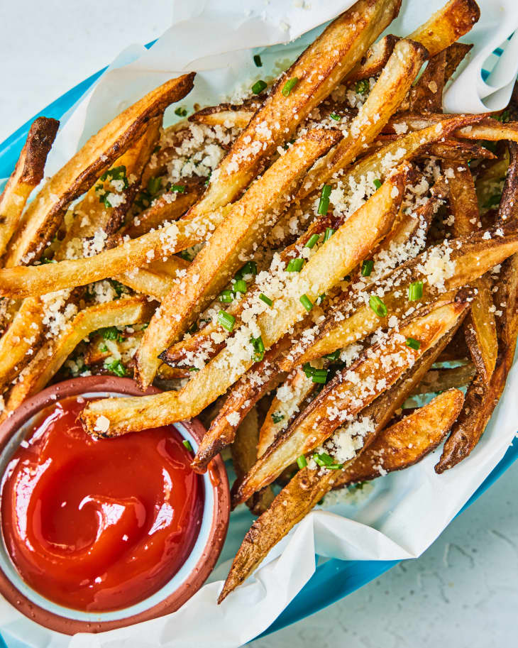 truffle french fries in a blue basket, with a white paper underneath, a side of ketchup, and white and green garnish on top.