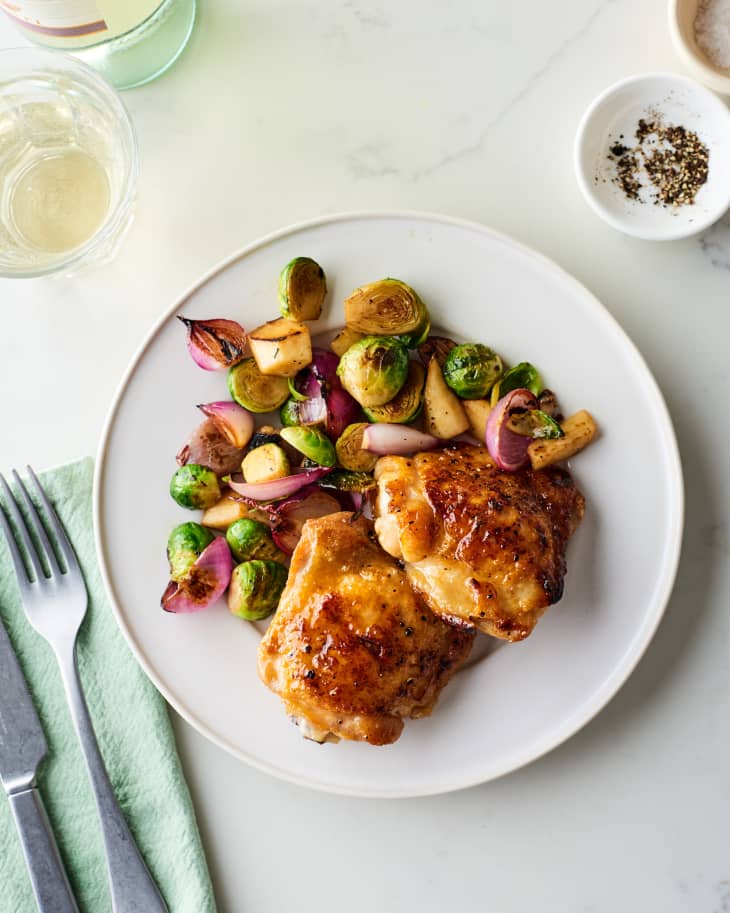 Roasted chicken thighs with Brussels sprouts, apples, and red onions on a white plate, next to a glass of white wine.