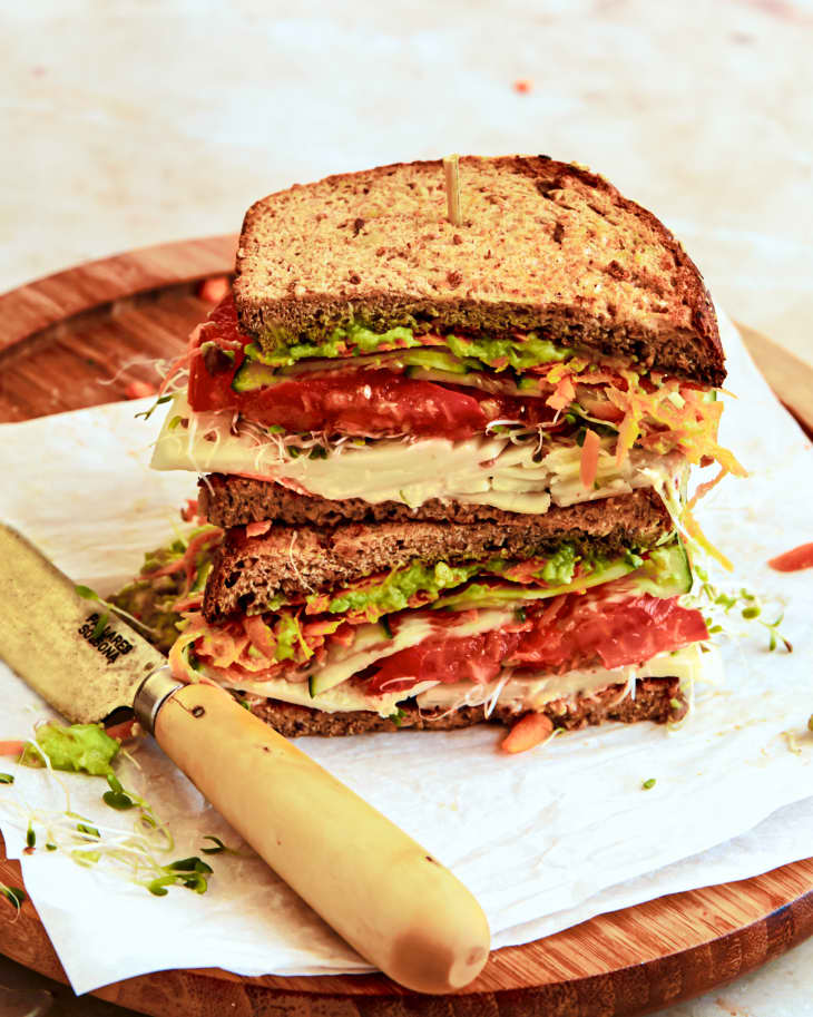 Stacked sandwich with whole grain bread, avocado, tomato, lettuce, and sprouts on a wooden board with a knife.