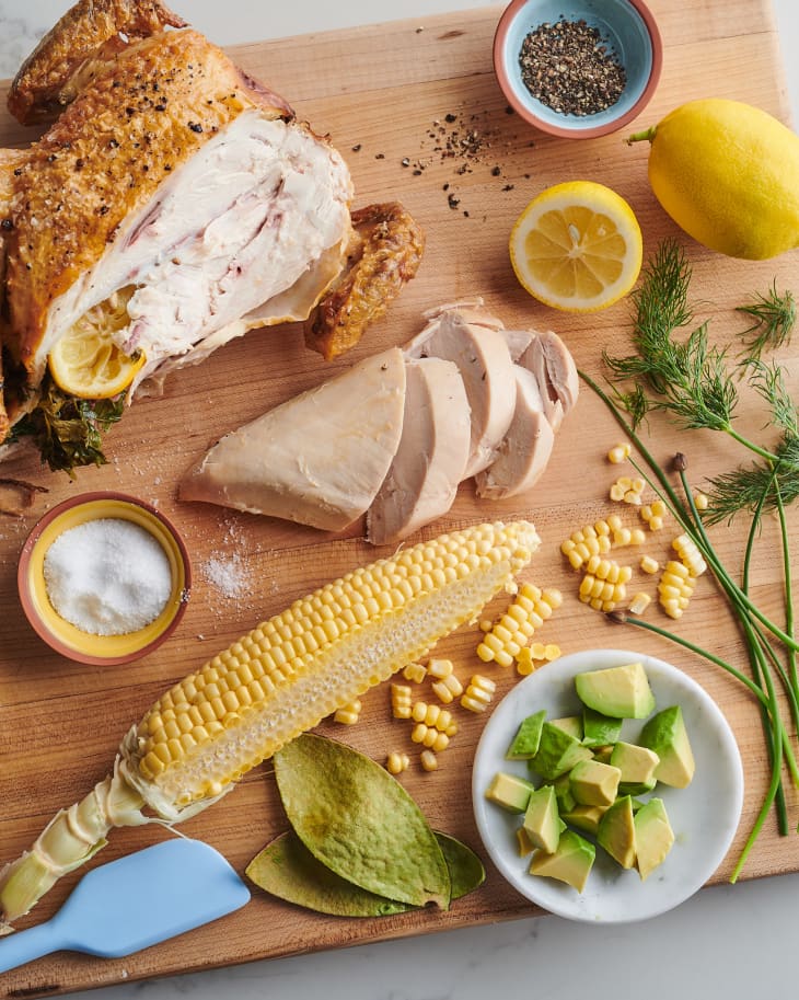 rotisserie chicken on cutting board next to bowl of salt, bowl of avocado, sliced lemon, and corn