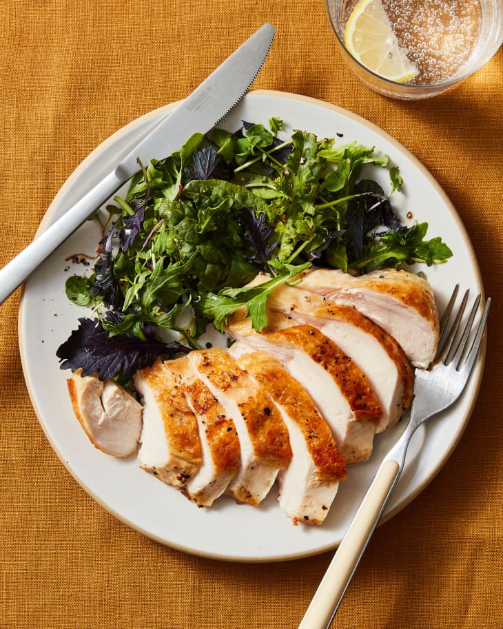 chicken breast prepared in a sous-vide, plated and sliced, and served with salad greens. A glass of seltzer with lemon in the upper right