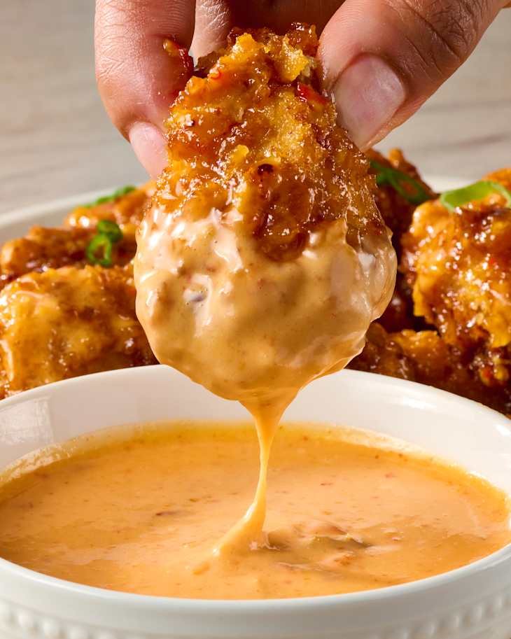 Crispy fried chicken piece being dipped into a creamy orange sauce, with green onions and more chicken pieces in the background.