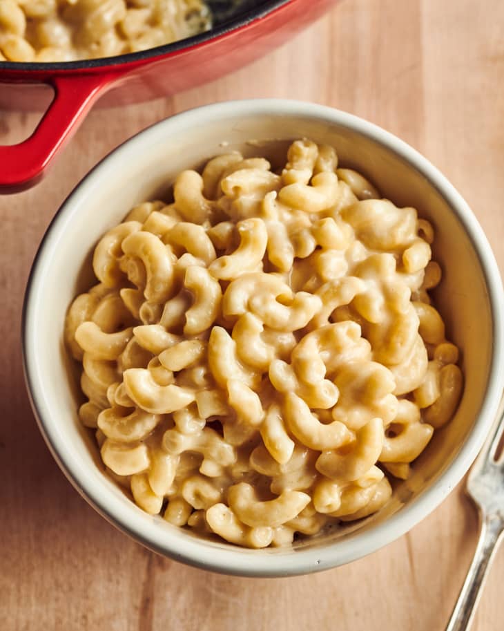 Creamy macaroni and cheese in a white bowl on a wooden table, with a red pot in the background.