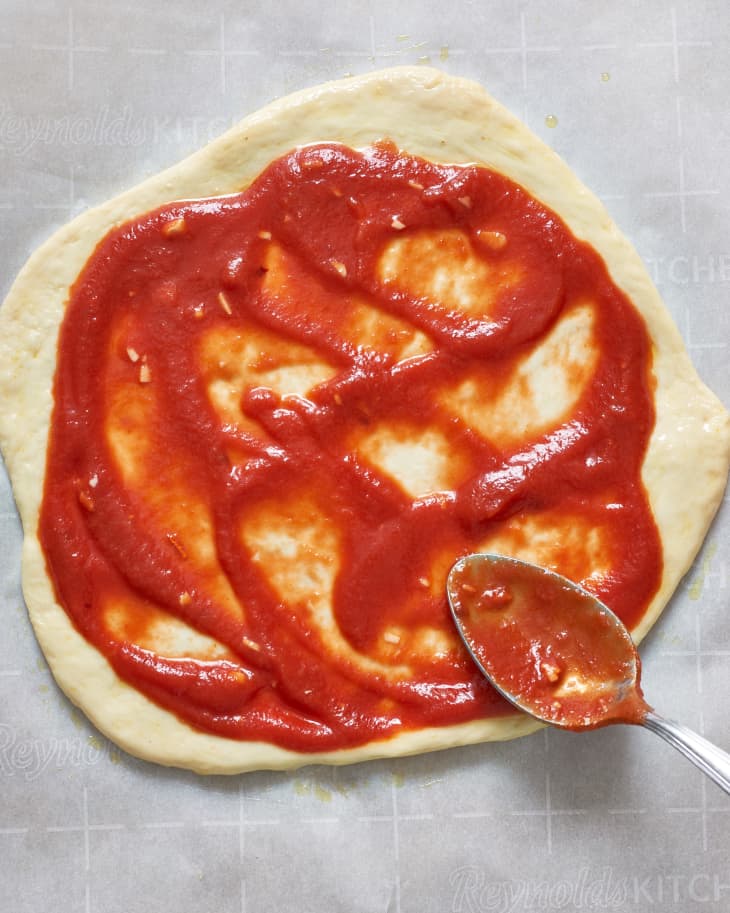 Pizza dough with tomato sauce spread by a spoon on parchment paper.