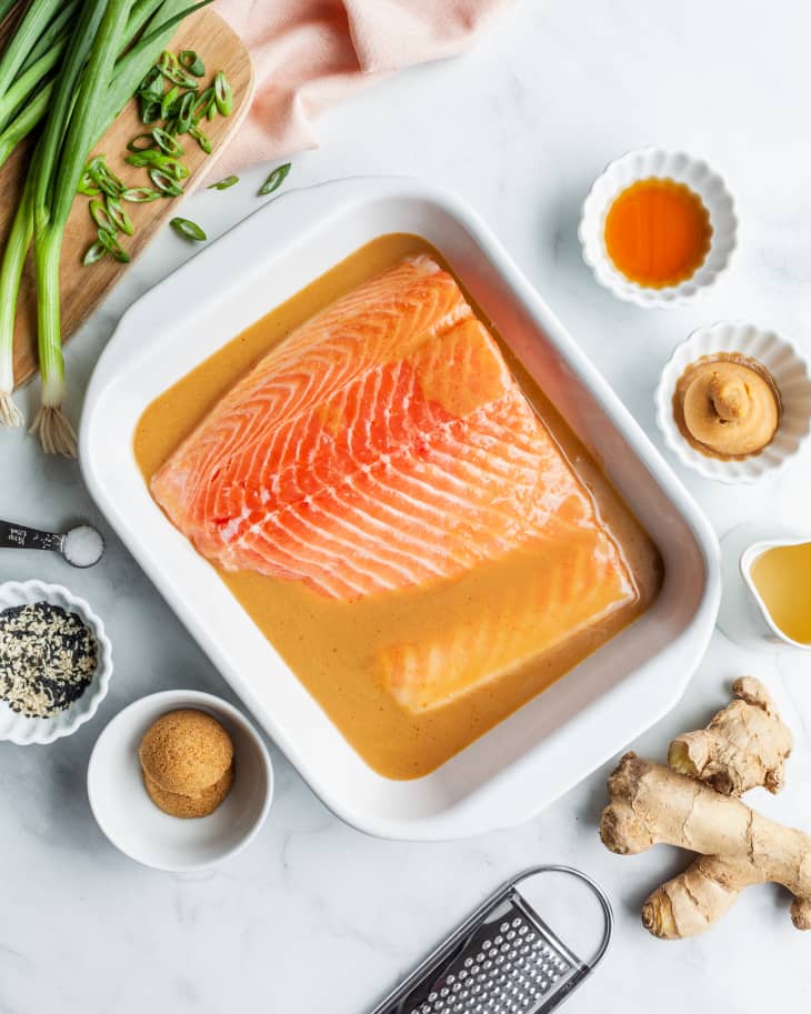 Salmon fillet in miso marinade with ginger, green onions, and sesame seeds on a white surface.
