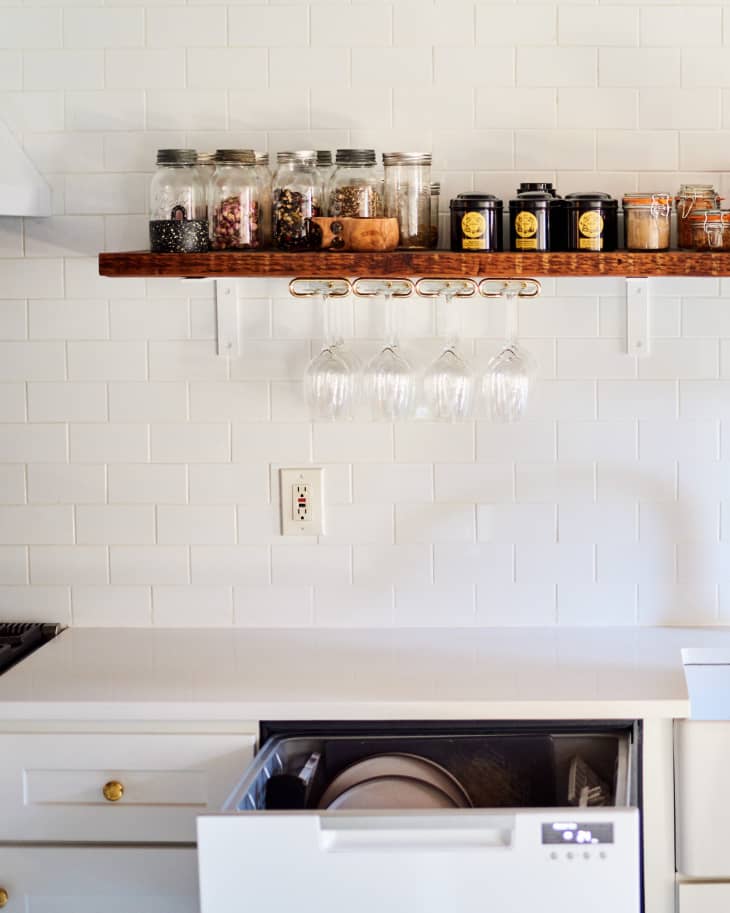 kitchen with white counters, white walls. Wood shelf with jars and canisters of food, wine glasses hanging underneath. Dishwasher below is open a bit and dishes can be seen