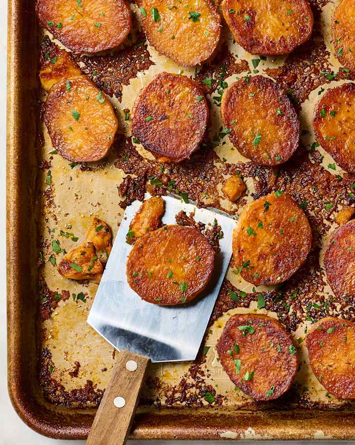 Crispy roasted potato slices garnished with parsley on a baking sheet, with a spatula resting on the side.