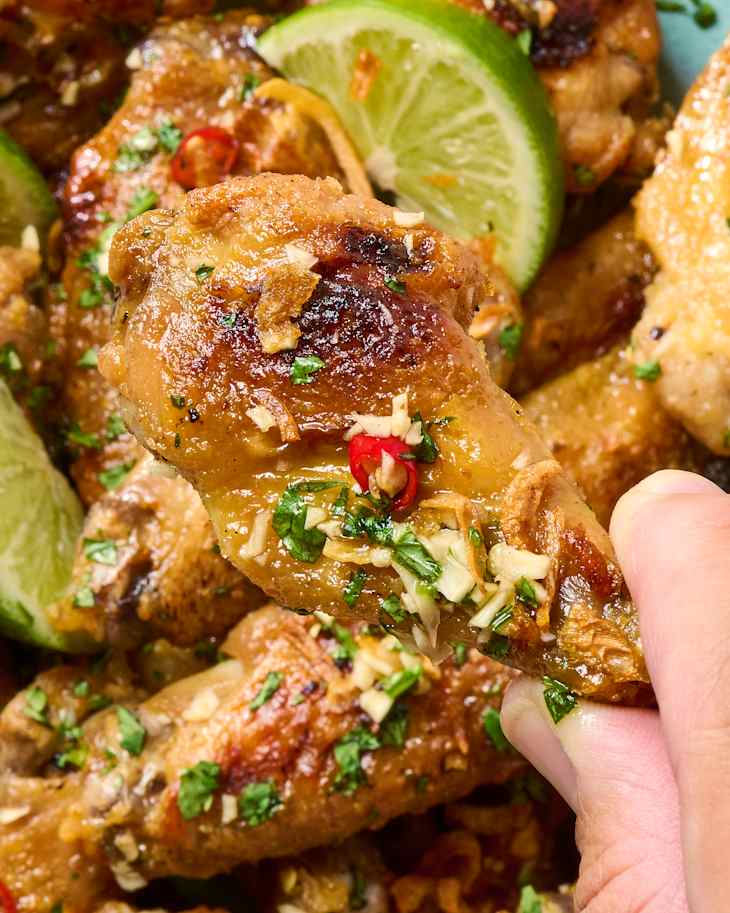 A hand holding a golden-brown chicken wing garnished with chopped herbs, garlic, and red chili, with a plate in the background.