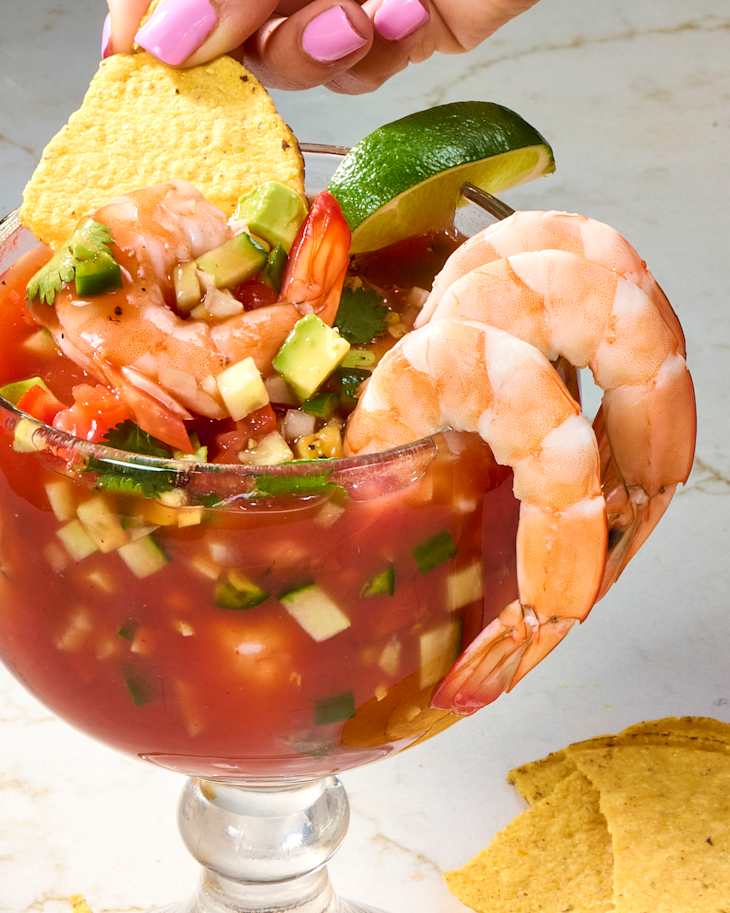 A glass bowl filled with shrimp cocktail, diced vegetables, and cilantro, garnished with lime and tortilla chips.