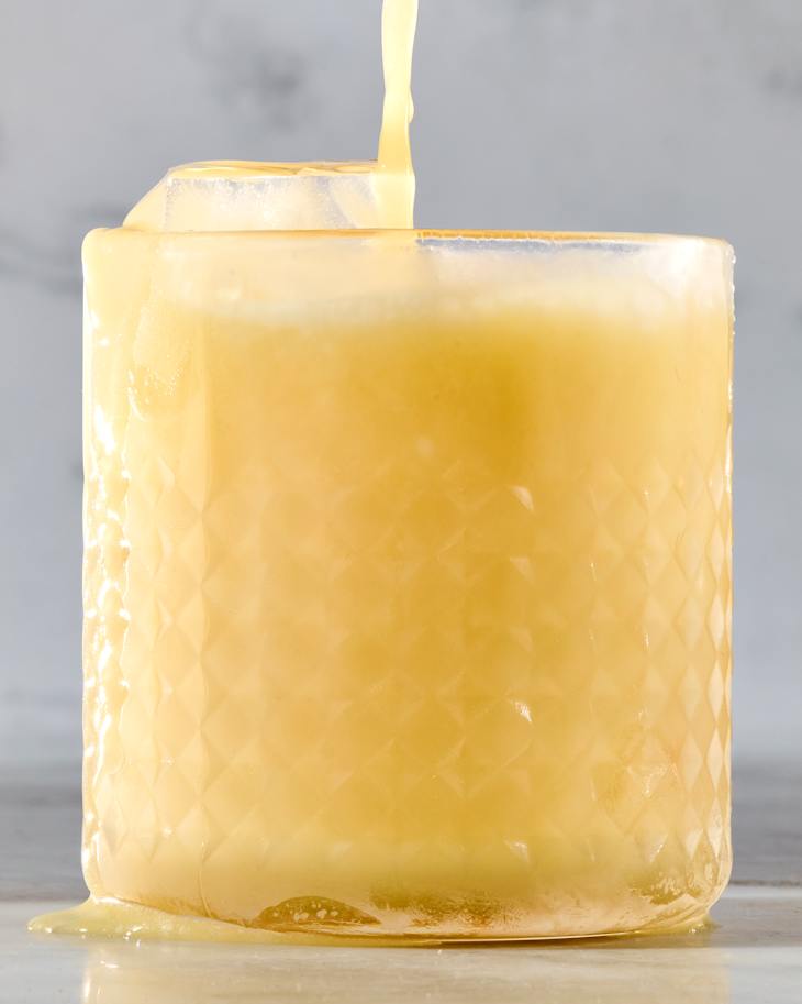 A textured glass filled with a yellow beverage, with liquid spilling over the rim.