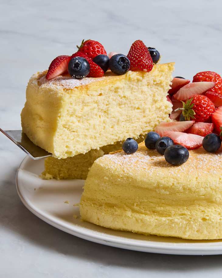 Fluffy cheesecake topped with strawberries and blueberries, served on a white plate with a slice being lifted.