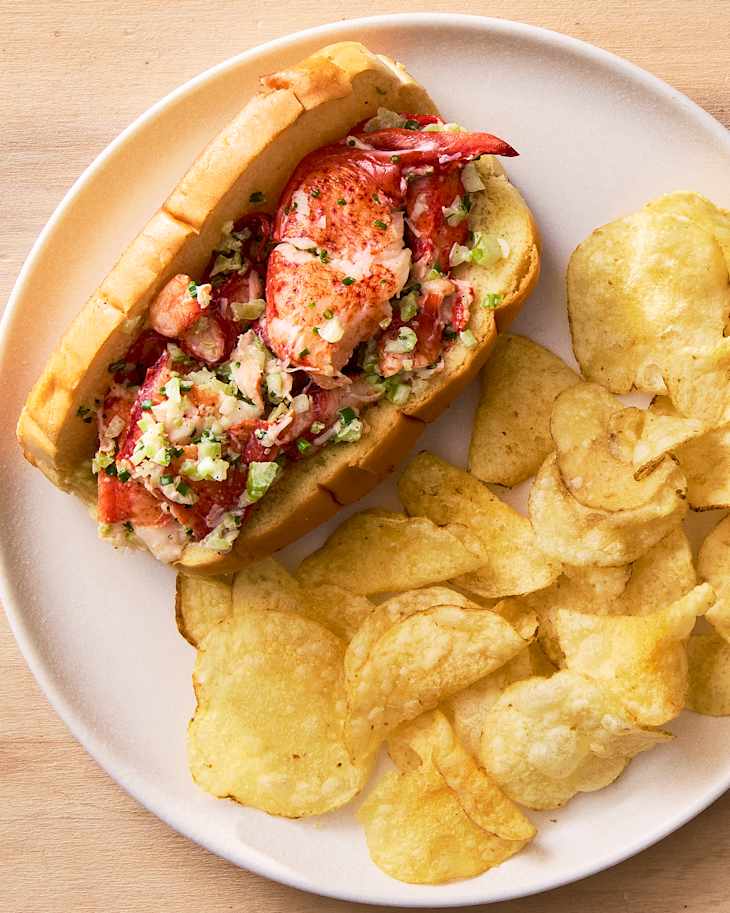 Lobster roll filled with lobster meat and chopped vegetables, served with a side of potato chips.