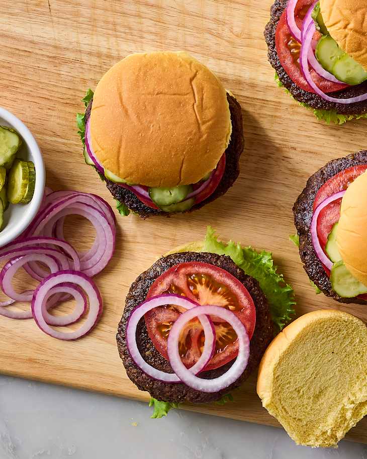 Four burgers on a wooden cutting board, topped with lettuce, tomato, pickles, and red onion, with a side of pickles.