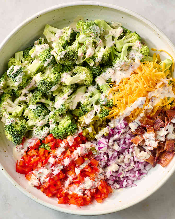 A large bowl filled with chopped broccoli, shredded cheese, diced tomatoes, red onions, and bacon, drizzled with dressing.