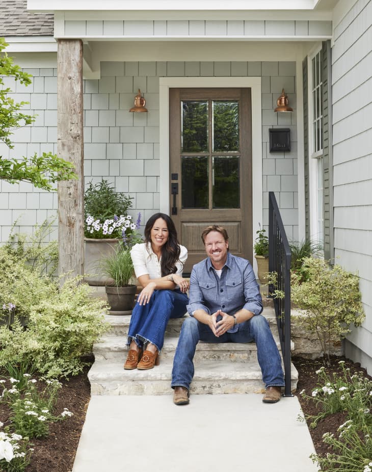 Couple sitting on stone steps of a gray shingle house with potted plants and copper light fixtures.