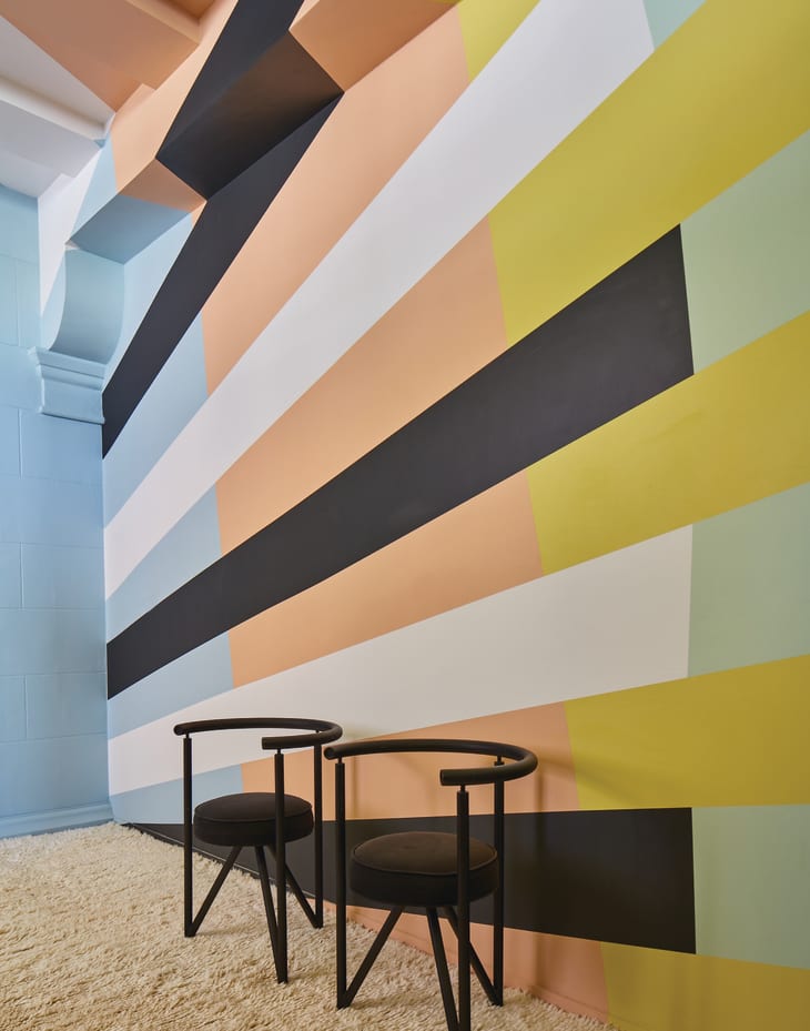 Two black chairs on a cream rug against a colorful geometric wall with stripes in pastel and bold colors.
