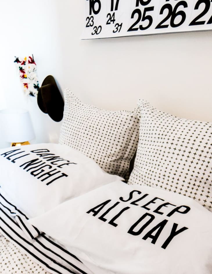 Bed with black and white patterned pillows, one reading "Dance All Night" and the other "Sleep All Day," under a calendar.