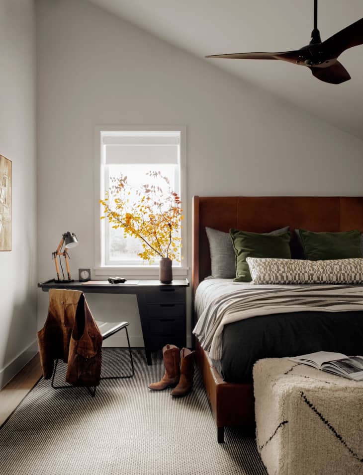 Cozy bedroom with a leather bed, green pillows, desk, chair, boots, and a window with autumn leaves.
