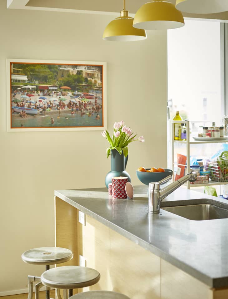 Kitchen with gray countertop, vase of pink tulips, fruit bowl, and colorful wall art under yellow pendant lights.