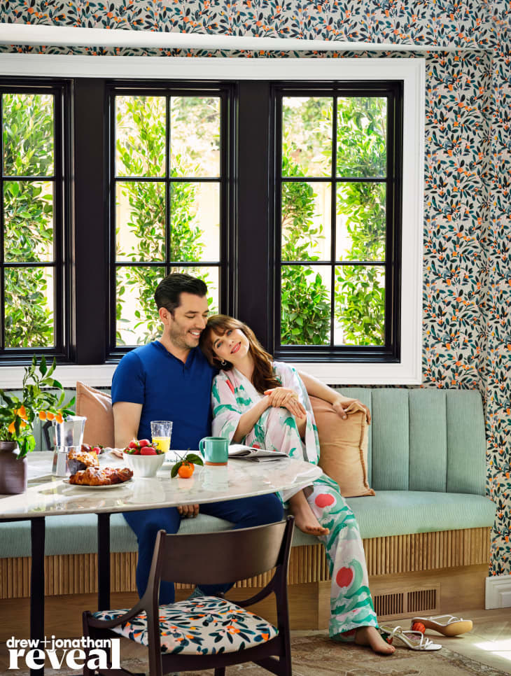 Couple sitting on a teal banquette in a floral wallpapered room, with a table set for breakfast.