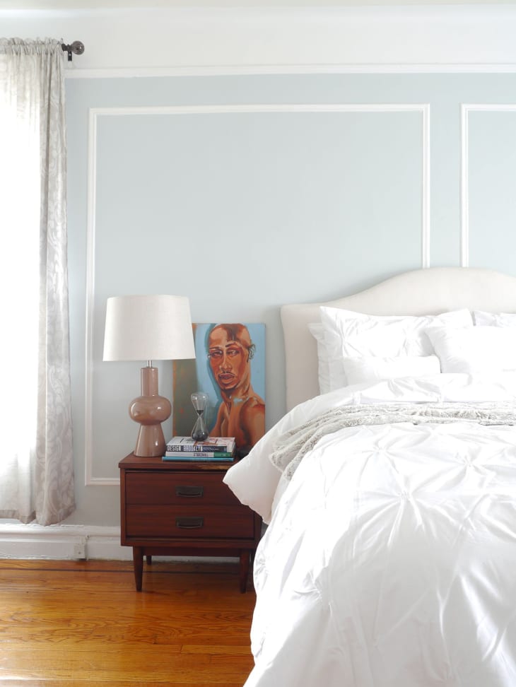 Bedroom with white bedding, wooden nightstand, lamp, books, and a portrait painting against a light blue wall.