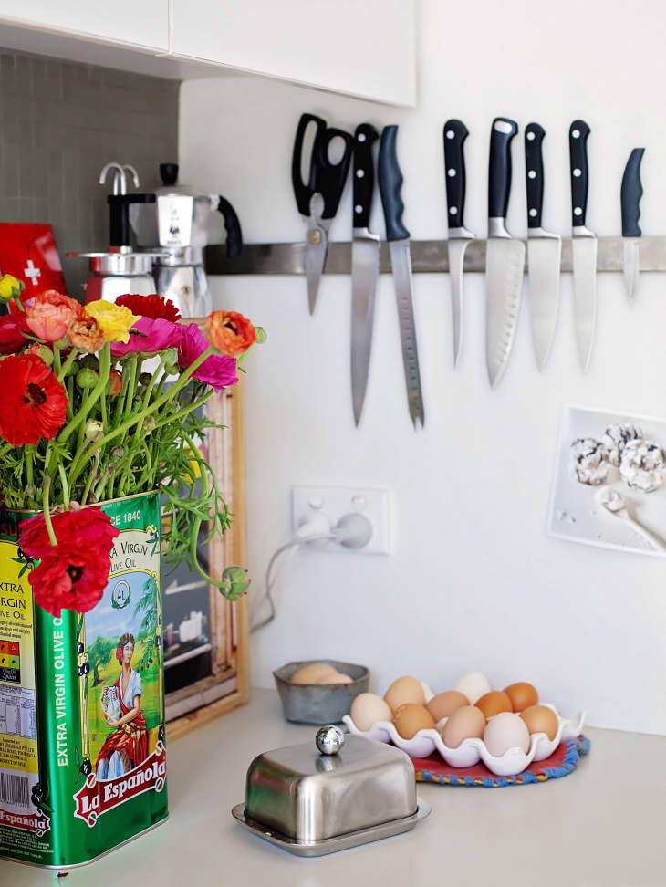 Colorful flowers in an olive oil tin, knives on a magnetic strip, eggs in a tray, and a butter dish on a kitchen counter.