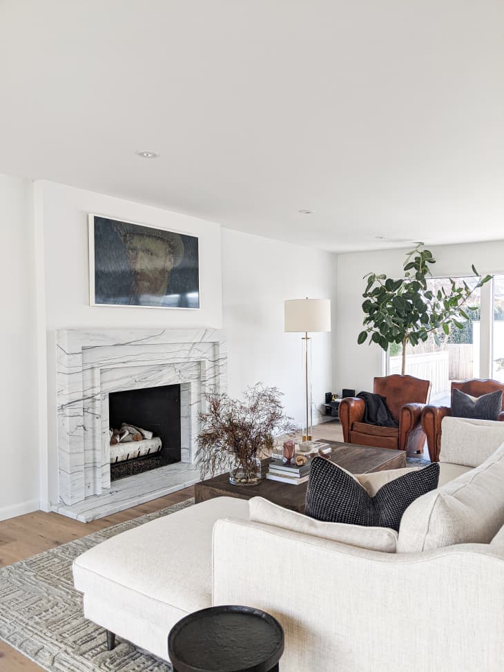 Living room with a marble fireplace, cream sectional sofa, leather armchair, floor lamp, and large potted plant.