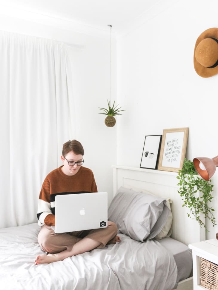 Person sitting on a bed with a laptop, surrounded by minimalist decor, including a hanging plant and framed art.