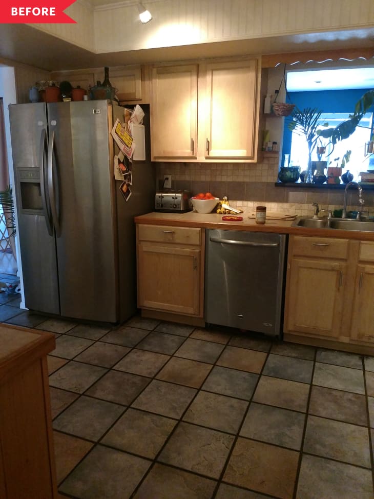 Kitchen with stainless steel fridge, light wood cabinets, tiled floor, and a window with plants.