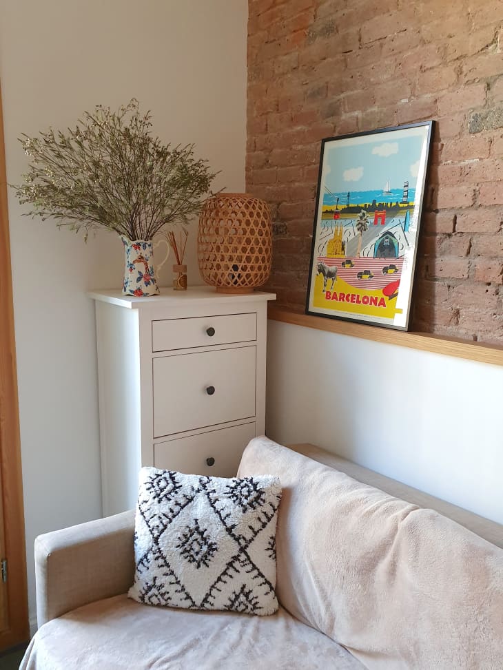 Cozy corner with beige sofa, patterned cushion, white dresser, floral vase, wicker lamp, and Barcelona poster on brick wall.