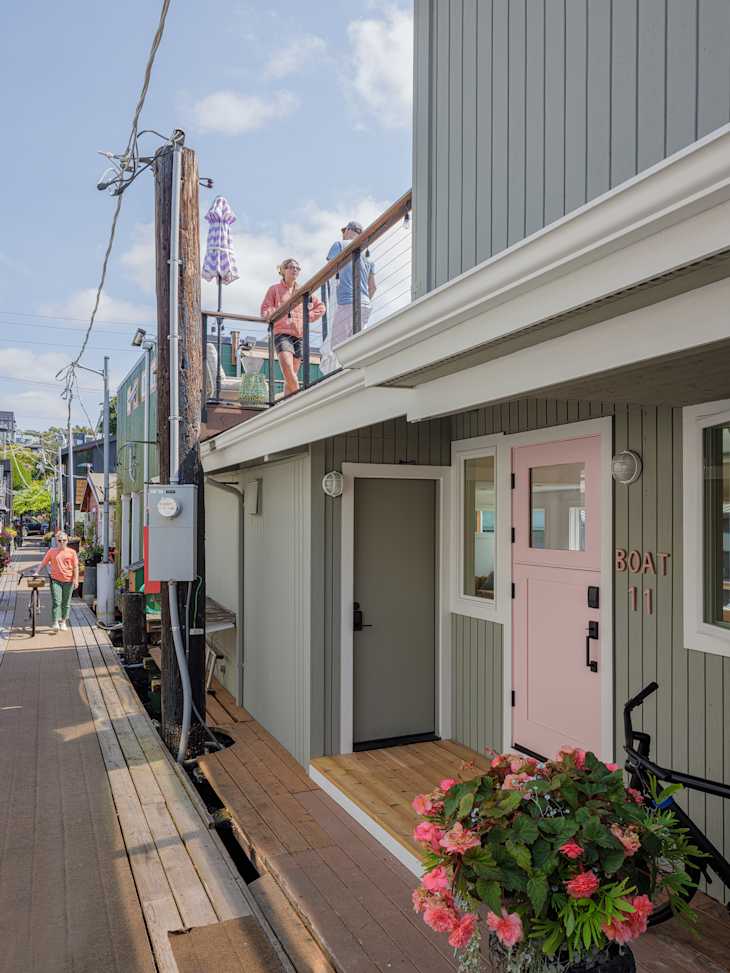 Two people on a balcony of a gray house, with a pink door labeled "BOAT 11," and a flower pot on the porch.
