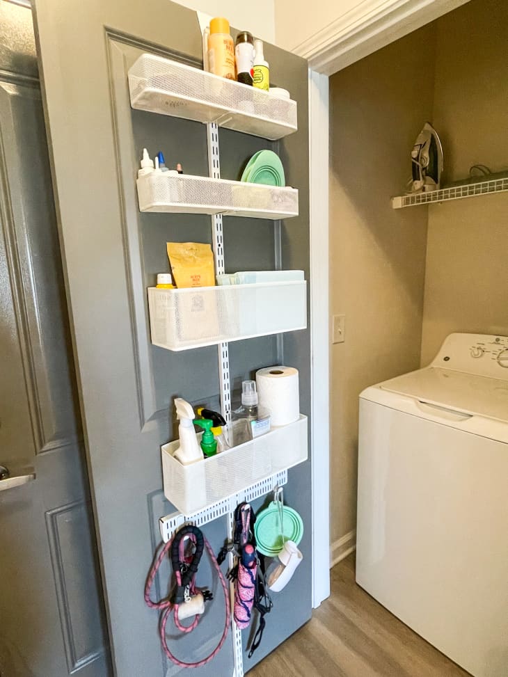 Over-the-door storage with cleaning supplies, paper towels, and dog leashes in a laundry room.